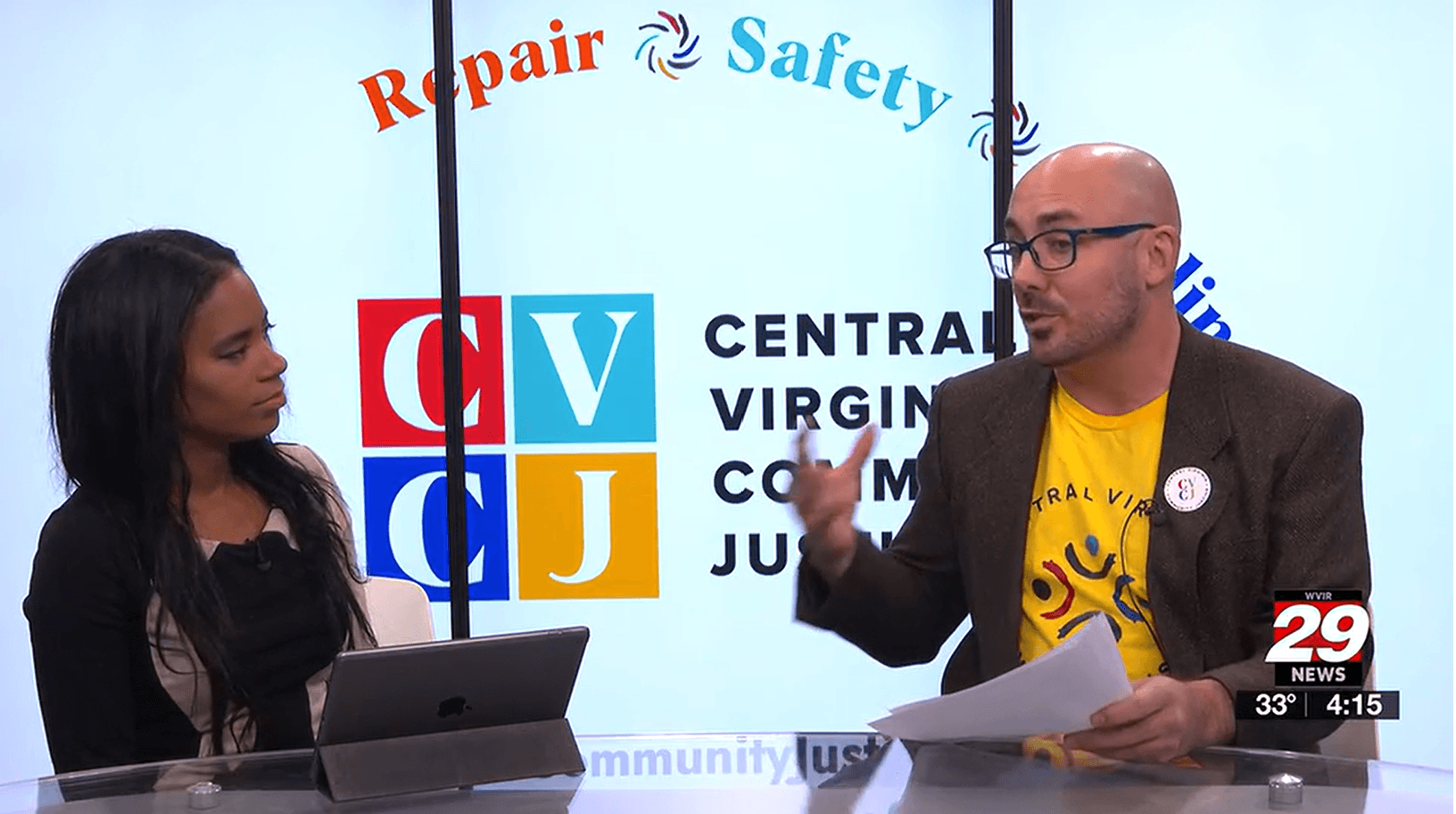 Woman and man speaking in front of a backdrop with CVCJ logo and the words Repair - Safety - Healing
